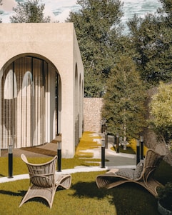 A modern architectural structure with large arching windows surrounded by a serene outdoor setting. The area features wicker chairs on a neatly trimmed lawn with stepping stones leading towards the building. Trees and shrubbery provide natural greenery, complementing the warm tones of the structure.