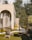 A modern architectural structure with large arching windows surrounded by a serene outdoor setting. The area features wicker chairs on a neatly trimmed lawn with stepping stones leading towards the building. Trees and shrubbery provide natural greenery, complementing the warm tones of the structure.