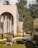A modern architectural structure with large arching windows surrounded by a serene outdoor setting. The area features wicker chairs on a neatly trimmed lawn with stepping stones leading towards the building. Trees and shrubbery provide natural greenery, complementing the warm tones of the structure.