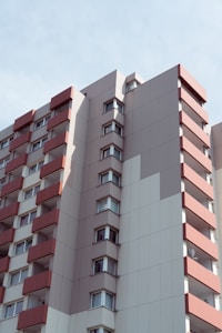 A modern high-rise apartment building with a minimalist design. The building features smooth surfaces with a beige and white color scheme, accented by red horizontal balcony lines. The windows are symmetrically arranged, creating a clean, orderly appearance. The sky is clear with subtle cloud patterns.