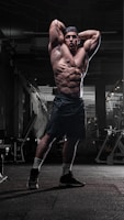 A muscular man is standing in a dimly lit gym with his arms raised above his head, showcasing his defined physique. The gym equipment surrounds him, and the lighting highlights his muscles and athletic build.