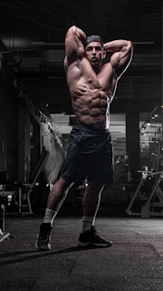 A muscular man is standing in a dimly lit gym with his arms raised above his head, showcasing his defined physique. The gym equipment surrounds him, and the lighting highlights his muscles and athletic build.