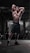 A muscular man is standing in a dimly lit gym with his arms raised above his head, showcasing his defined physique. The gym equipment surrounds him, and the lighting highlights his muscles and athletic build.