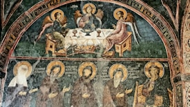 A religious fresco with angelic figures seated around a table, possibly a depiction of the Holy Trinity, surrounded by additional haloed figures below. The painting is rich in detail and set against a dark green background, depicting traditional religious iconography with a medieval artistic style.