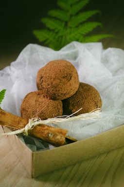 A small box contains several chocolate truffles dusted with cocoa powder, resting on a bed of white fabric. A cinnamon stick is tied with a string, adding a rustic touch. There are green fern leaves in the background providing a natural, earthy ambiance.