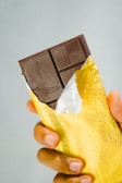 A hand holding a partially unwrapped dark chocolate bar with a gold foil wrapper. The chocolate bar has intricate designs etched on the surface.