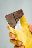 A hand holding a partially unwrapped dark chocolate bar with a gold foil wrapper. The chocolate bar has intricate designs etched on the surface.