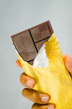 A hand holding a partially unwrapped dark chocolate bar with a gold foil wrapper. The chocolate bar has intricate designs etched on the surface.