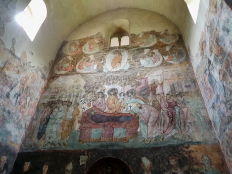 A detailed fresco depicting a religious scene inside a church, featuring a central figure surrounded by several other figures, some with halos. The mural shows people in flowing robes, some holding crosses, and other religious symbols. The scene is painted on a curved wall beneath a small arched window, with additional smaller scenes and figures filling the space above.