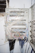 A metal electrical panel is filled with numerous wires of varying colors, including white, blue, red, and yellow. The wires are organized through several rows of terminal blocks within the panel. The panel is mounted on a wall with exposed plaster and nearby wire bundles are visible.