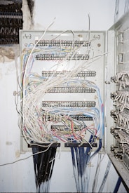 A metal electrical panel is filled with numerous wires of varying colors, including white, blue, red, and yellow. The wires are organized through several rows of terminal blocks within the panel. The panel is mounted on a wall with exposed plaster and nearby wire bundles are visible.