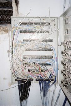 A metal electrical panel is filled with numerous wires of varying colors, including white, blue, red, and yellow. The wires are organized through several rows of terminal blocks within the panel. The panel is mounted on a wall with exposed plaster and nearby wire bundles are visible.