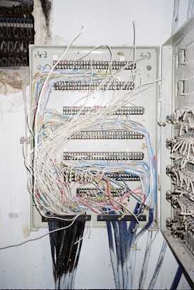 A metal electrical panel is filled with numerous wires of varying colors, including white, blue, red, and yellow. The wires are organized through several rows of terminal blocks within the panel. The panel is mounted on a wall with exposed plaster and nearby wire bundles are visible.