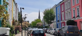 A street view featuring a row of colorful houses with distinctly painted facades of pastel shades, including blue, pink, and yellow. Lush greenery and flowering plants enhance the sidewalk, while parked cars line the street. A pointed church steeple is visible in the background beneath a partly cloudy sky.