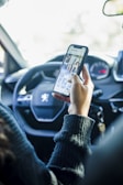A person holding a smartphone in one hand inside a car, with the phone screen displaying a social media app. The steering wheel, car dashboard, and a set of keys are visible.