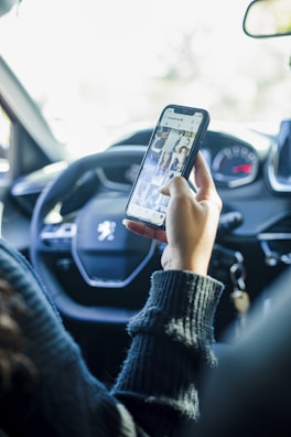 A person holding a smartphone in one hand inside a car, with the phone screen displaying a social media app. The steering wheel, car dashboard, and a set of keys are visible.