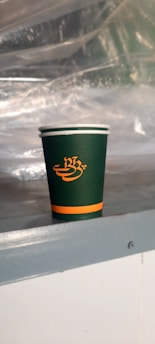 A green disposable cup with an orange logo and stripe, resting on a metallic surface. In the background, there is a transparent plastic sheet creating a blurred effect.