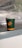 A green disposable cup with an orange logo and stripe, resting on a metallic surface. In the background, there is a transparent plastic sheet creating a blurred effect.
