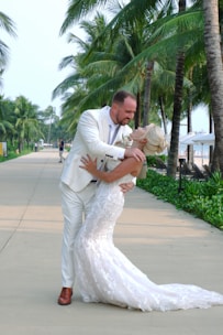 A couple in formal attire embraces joyfully beneath tall, lush palm trees on a sunlit pathway. The groom is dressed in a white suit with brown shoes, while the bride wears a long, elegant white lace gown. The scene conveys a sense of celebration and romance.