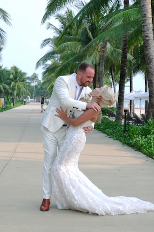 A couple in formal attire embraces joyfully beneath tall, lush palm trees on a sunlit pathway. The groom is dressed in a white suit with brown shoes, while the bride wears a long, elegant white lace gown. The scene conveys a sense of celebration and romance.