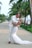 A couple in formal attire embraces joyfully beneath tall, lush palm trees on a sunlit pathway. The groom is dressed in a white suit with brown shoes, while the bride wears a long, elegant white lace gown. The scene conveys a sense of celebration and romance.
