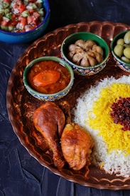 A beautifully arranged platter includes cooked chicken drumsticks, rice topped with saffron and barberries, accompanied by small bowls of soup with a carrot slice, garlic, and olives. The set is presented on a decorative copper tray. In the background, a bowl containing a fresh salad of tomatoes, cucumbers, and herbs adds a colorful contrast.