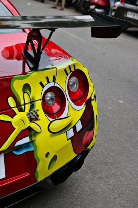 A close-up of a car's rear end featuring a prominent custom graphic of a cartoon character covering the vehicle's fender and bumper. The design is bright and playful, with large red eyes and a big smile, incorporating the car's tail lights as part of the character's features. The red car has a large rear spoiler and is parked on a street with other vehicles in the background.