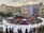 An open-air event is taking place in front of a shopping mall with branded stores. A stage is set up for performances, surrounded by a crowd of people. White tents are positioned around the area, possibly for vendors or information booths. There's an inflatable game setup that appears to be a mechanical bull ride. The atmosphere seems lively and crowded.