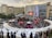 An open-air event is taking place in front of a shopping mall with branded stores. A stage is set up for performances, surrounded by a crowd of people. White tents are positioned around the area, possibly for vendors or information booths. There's an inflatable game setup that appears to be a mechanical bull ride. The atmosphere seems lively and crowded.