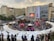 An open-air event is taking place in front of a shopping mall with branded stores. A stage is set up for performances, surrounded by a crowd of people. White tents are positioned around the area, possibly for vendors or information booths. There's an inflatable game setup that appears to be a mechanical bull ride. The atmosphere seems lively and crowded.