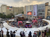 An open-air event is taking place in front of a shopping mall with branded stores. A stage is set up for performances, surrounded by a crowd of people. White tents are positioned around the area, possibly for vendors or information booths. There's an inflatable game setup that appears to be a mechanical bull ride. The atmosphere seems lively and crowded.