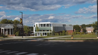 A modern, glass-fronted building with multiple stories sits on a well-maintained grassy area surrounded by trees. Traditional brick buildings with columns are nearby, indicating a campus or institutional setting. The sky is partly cloudy, creating a serene atmosphere.