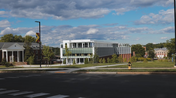 A modern, glass-fronted building with multiple stories sits on a well-maintained grassy area surrounded by trees. Traditional brick buildings with columns are nearby, indicating a campus or institutional setting. The sky is partly cloudy, creating a serene atmosphere.