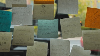 Several colorful sticky notes are attached to a glass surface, each with various handwritten messages and drawings. The notes are in different colors including brown, blue, and orange, featuring motivational quotes and simple illustrations like smiley faces and stick figures.