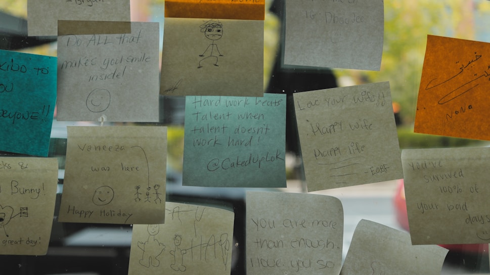 Several colorful sticky notes are attached to a glass surface, each with various handwritten messages and drawings. The notes are in different colors including brown, blue, and orange, featuring motivational quotes and simple illustrations like smiley faces and stick figures.