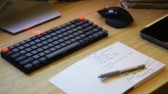 A neatly arranged workspace featuring a sleek keyboard with orange accents, a wireless mouse, a notebook with handwritten notes, and a pen resting on it. There is also part of a laptop visible, suggesting a tech-focused setting.