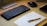 A neatly arranged workspace featuring a sleek keyboard with orange accents, a wireless mouse, a notebook with handwritten notes, and a pen resting on it. There is also part of a laptop visible, suggesting a tech-focused setting.