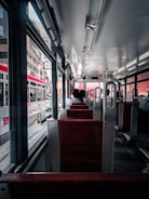 A view from inside a public transit vehicle with wooden seats and metal frames. A few passengers are seated, facing forward. The vehicle is traveling through an urban area, visible through large windows, with several buildings and another similar transit vehicle on the adjacent track. The lighting is natural, coming from both outside and overhead lights.