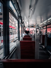 A view from inside a public transit vehicle with wooden seats and metal frames. A few passengers are seated, facing forward. The vehicle is traveling through an urban area, visible through large windows, with several buildings and another similar transit vehicle on the adjacent track. The lighting is natural, coming from both outside and overhead lights.