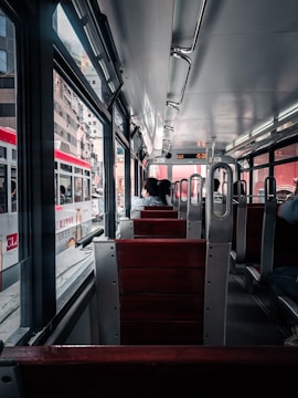 A view from inside a public transit vehicle with wooden seats and metal frames. A few passengers are seated, facing forward. The vehicle is traveling through an urban area, visible through large windows, with several buildings and another similar transit vehicle on the adjacent track. The lighting is natural, coming from both outside and overhead lights.