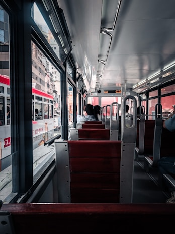 A view from inside a public transit vehicle with wooden seats and metal frames. A few passengers are seated, facing forward. The vehicle is traveling through an urban area, visible through large windows, with several buildings and another similar transit vehicle on the adjacent track. The lighting is natural, coming from both outside and overhead lights.