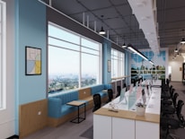A modern open-plan office space featuring a long row of desks with computers and chairs on a polished wooden floor. Large windows allow natural light to flood the room, showing a scenic cityscape outside. The walls are painted a light blue with decorative artwork and a vibrant mural depicting a city skyline. Comfortable blue seating and small tables are arranged along one side of the windows.