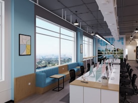 A modern open-plan office space featuring a long row of desks with computers and chairs on a polished wooden floor. Large windows allow natural light to flood the room, showing a scenic cityscape outside. The walls are painted a light blue with decorative artwork and a vibrant mural depicting a city skyline. Comfortable blue seating and small tables are arranged along one side of the windows.