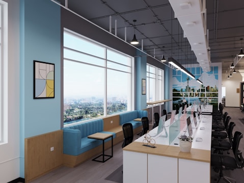 A modern open-plan office space featuring a long row of desks with computers and chairs on a polished wooden floor. Large windows allow natural light to flood the room, showing a scenic cityscape outside. The walls are painted a light blue with decorative artwork and a vibrant mural depicting a city skyline. Comfortable blue seating and small tables are arranged along one side of the windows.