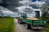 A green tractor with a covered trailer full of grain is on a narrow country road surrounded by grassy fields. Clouds in varying shades of gray fill the sky, creating an overcast atmosphere. To the right, there is a wooden hunting stand among thick bushes and trees.