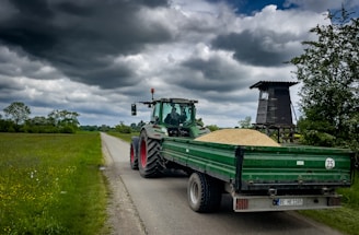 A green tractor with a covered trailer full of grain is on a narrow country road surrounded by grassy fields. Clouds in varying shades of gray fill the sky, creating an overcast atmosphere. To the right, there is a wooden hunting stand among thick bushes and trees.