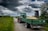 A green tractor with a covered trailer full of grain is on a narrow country road surrounded by grassy fields. Clouds in varying shades of gray fill the sky, creating an overcast atmosphere. To the right, there is a wooden hunting stand among thick bushes and trees.