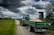 A green tractor with a covered trailer full of grain is on a narrow country road surrounded by grassy fields. Clouds in varying shades of gray fill the sky, creating an overcast atmosphere. To the right, there is a wooden hunting stand among thick bushes and trees.