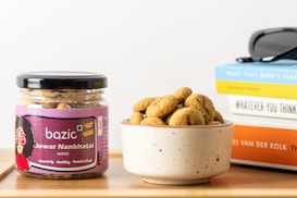 A jar labeled 'bazic Jowar Nankhatai Minis' is placed next to a small bowl filled with round cookies. The jar has a purple label and indicates gluten-free and natural sugars. Behind them, a stack of books with visible titles sits on the wooden surface, along with a pair of sunglasses.