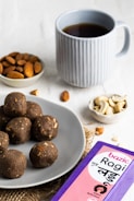 A light grey plate contains several dark brown, round snacks, possibly ragi laddus with visible nut pieces. Nearby, a small bowl of almonds and another with cashew nuts are placed. A blue mug filled with a dark beverage, possibly tea or coffee, is positioned in the background. A purple and pink box labeled 'Bazic Ragi Ladu' is visible in the foreground.